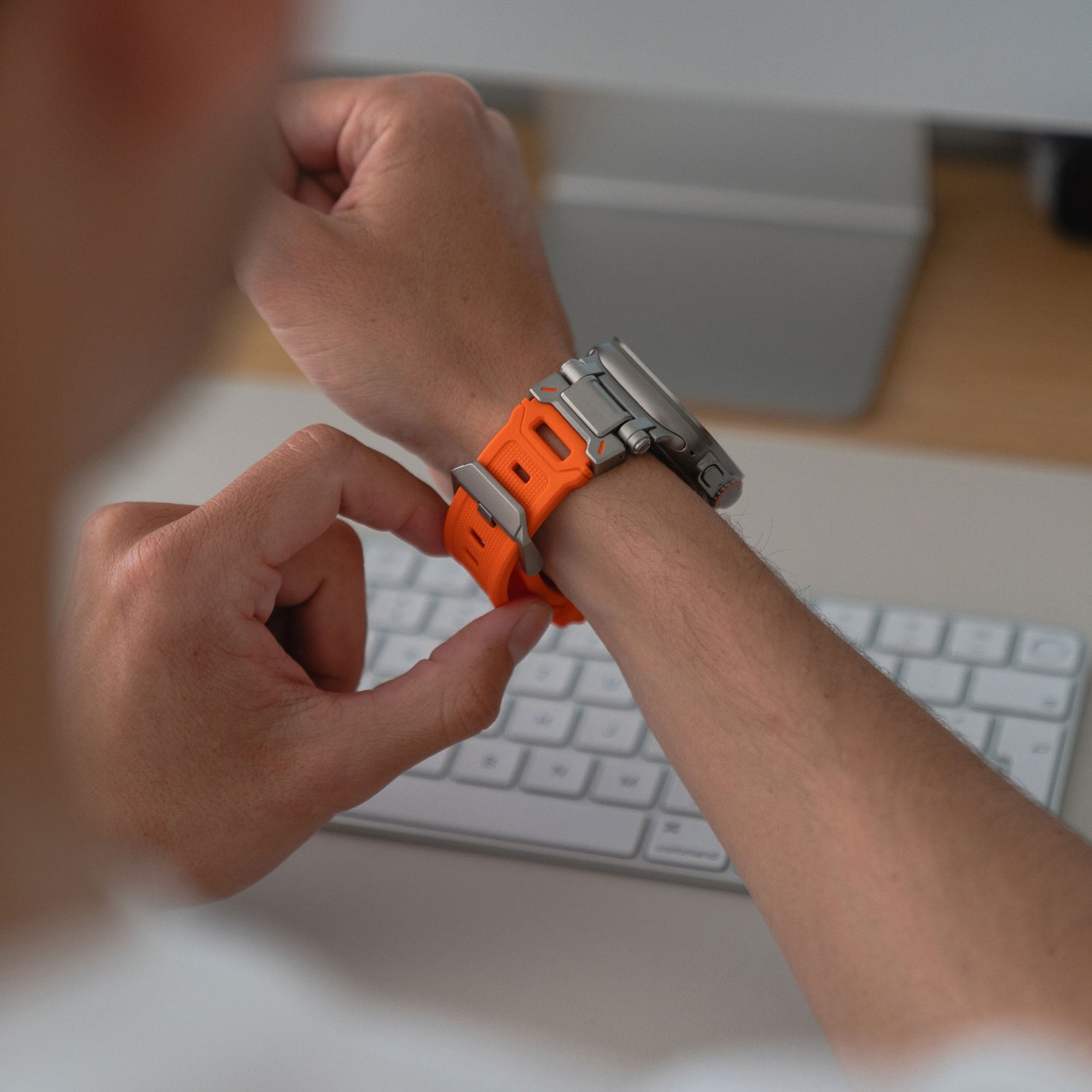 Eine Person befestigt das arktisband Apple Watch Ultra Armband „Atlas“ von arktisband an ihrem linken Handgelenk an einem Schreibtisch mit einer weißen Tastatur und einem Monitor im Hintergrund.