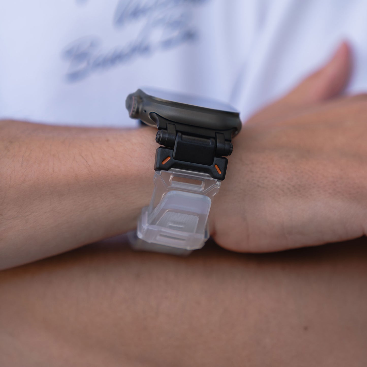 Eine Nahaufnahme der verschränkten Arme einer Person zeigt das Arktisband Apple Watch Ultra Armband „Atlas“ mit transparentem Armband und schwarzem Gehäuse mit orangefarbenen Akzenten, getragen über einem weißen Hemd.
