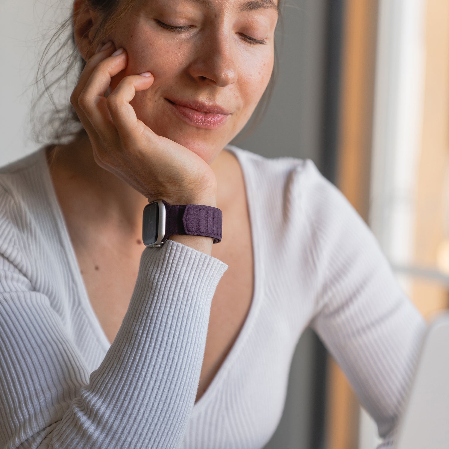Eine Frau mit geschlossenen Augen lächelt sanft und stützt ihr Kinn auf ihre Hand, während sie das elegante Arktisband Apple Watch magnetisches Armband aus Alcantara trägt.