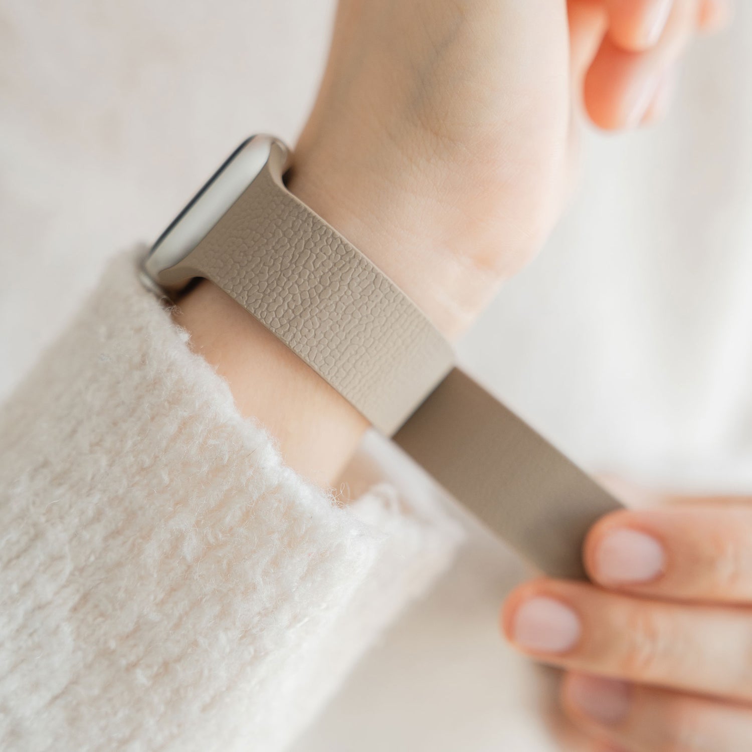 Nahaufnahme einer Person, die das Arktisband Apple Watch magnetisches Silikonarmband "Flexy" in Beige an ihrem Handgelenk befestigt, gepaart mit einem weichen Pullover.