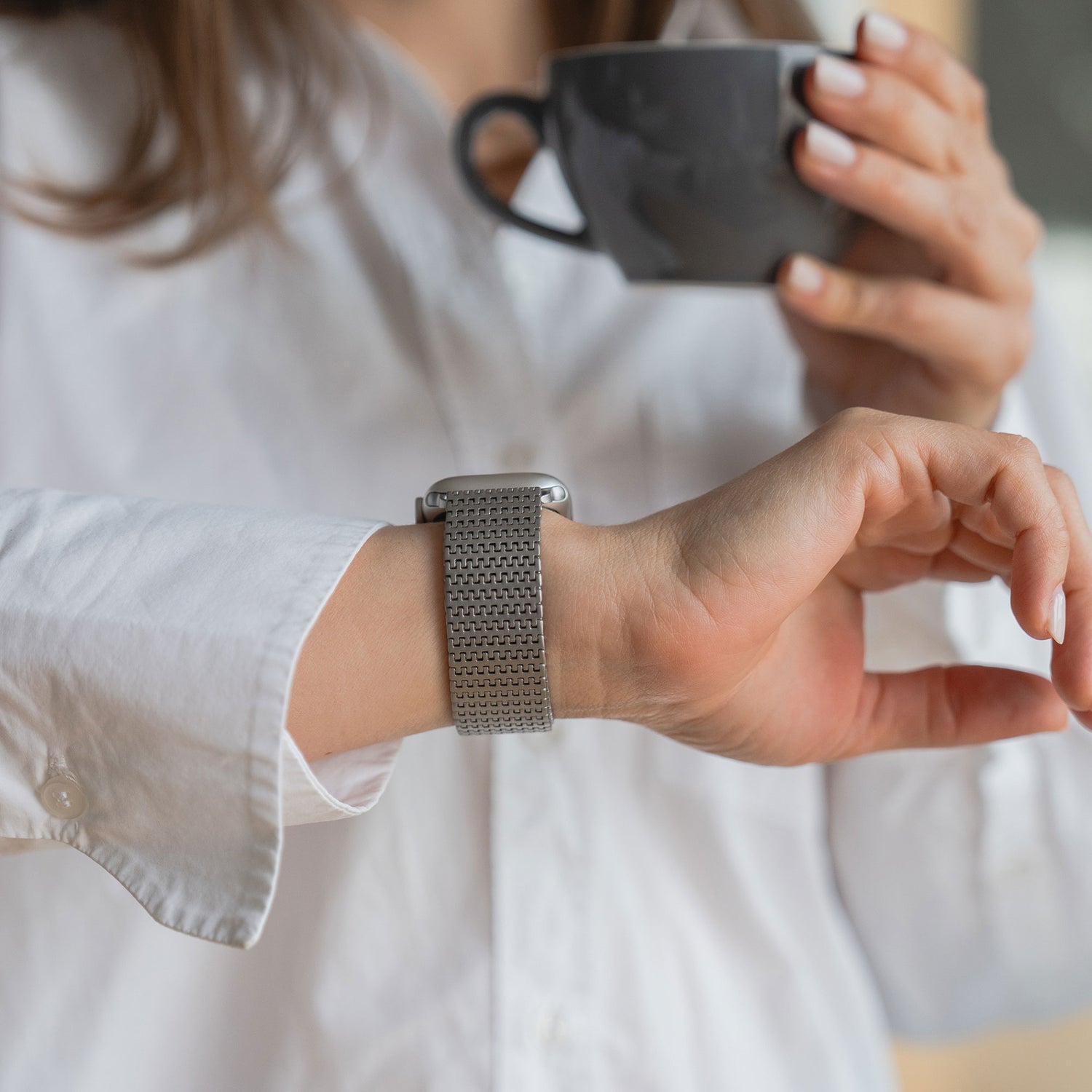 Eine Person im weißen Hemd hält eine dunkle Tasse und checkt die Uhrzeit auf ihrem Arktisband Apple Watch Edelstahlarmband „Aurora“.