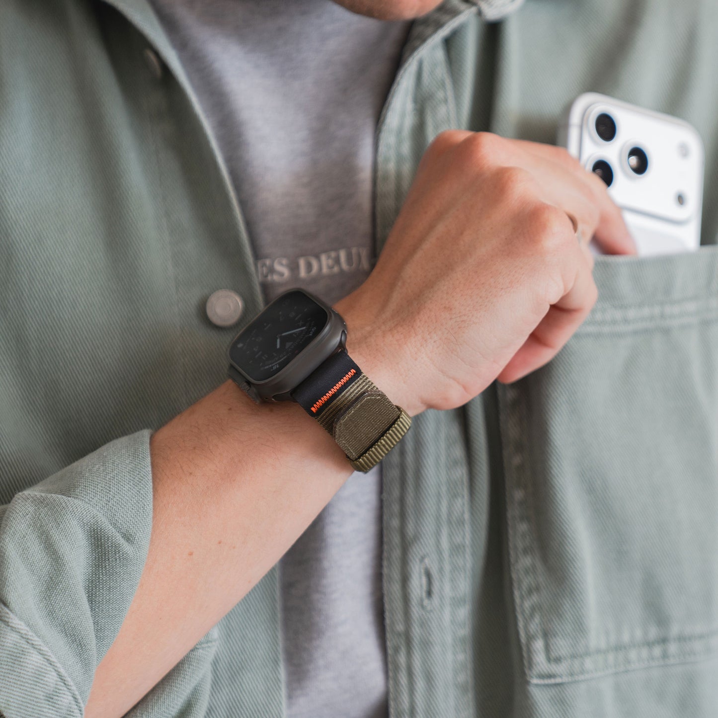 Eine Person trägt das arktisband Apple Watch Armband "Terra" von arktisband an ihrer Apple Watch Ultra und steckt ein weißes Smartphone in die Brusttasche eines grünen Hemdes.
