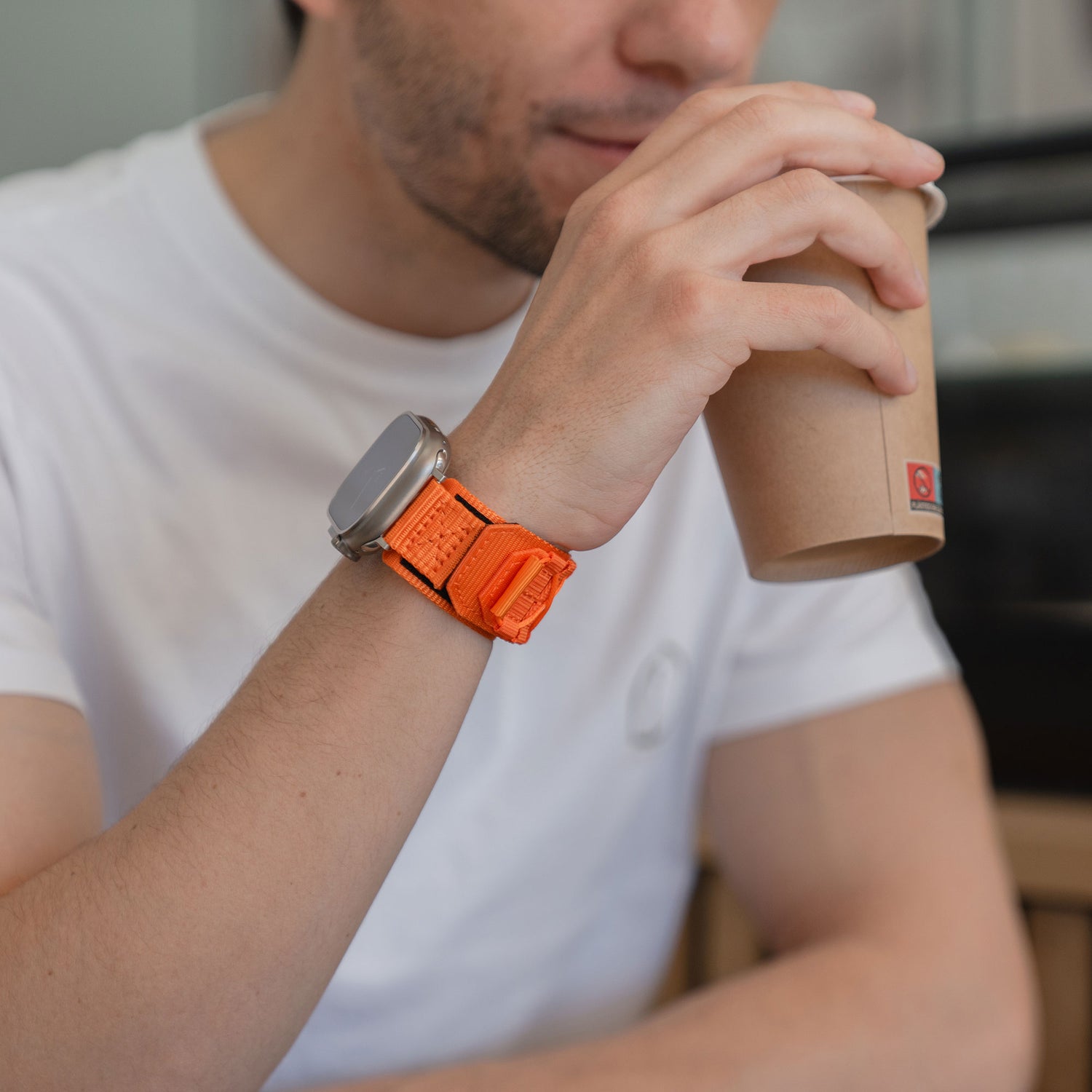 Ein Mann in einem weißen T-Shirt trinkt aus einem Pappbecher, während er das arktisband Apple Watch Outdoor Armband "Forge" mit einem orangefarbenen Nylonband trägt.