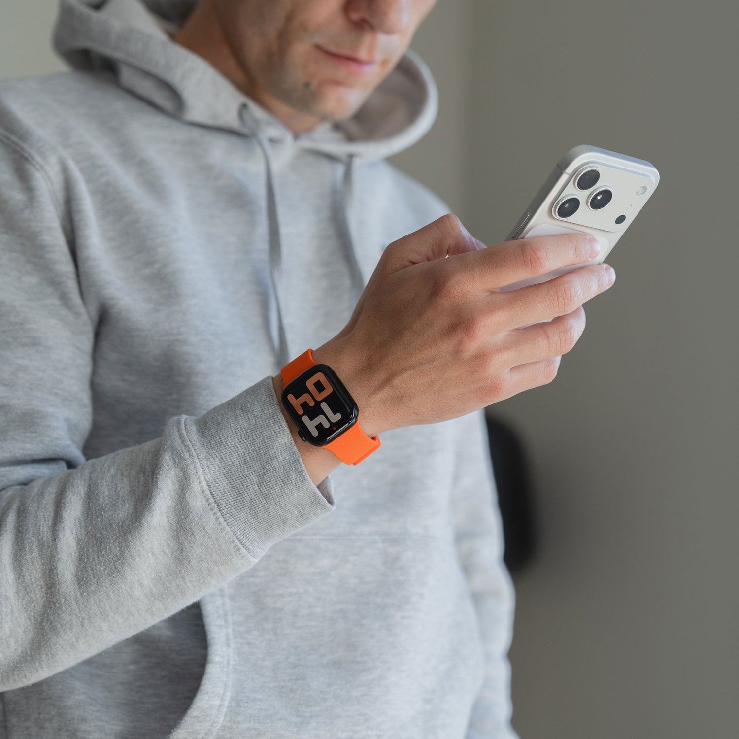Mann im grauen Kapuzenpulli, der ein Smartphone benutzt, trägt ein orangefarbenes Arktisband Apple Watch magnetisches Silikonarmband "Flexy" mit HAL auf dem Bildschirm.