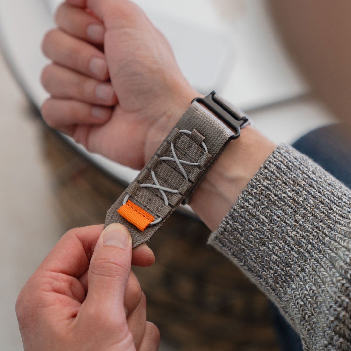 Jemand legt sich das arktisband Apple Watch Outdoor Armband "Forge", ein graues Nylonarmband mit weißen Schnürsenkeln und einem orangefarbenen Akzent von arktisband, um das Handgelenk.