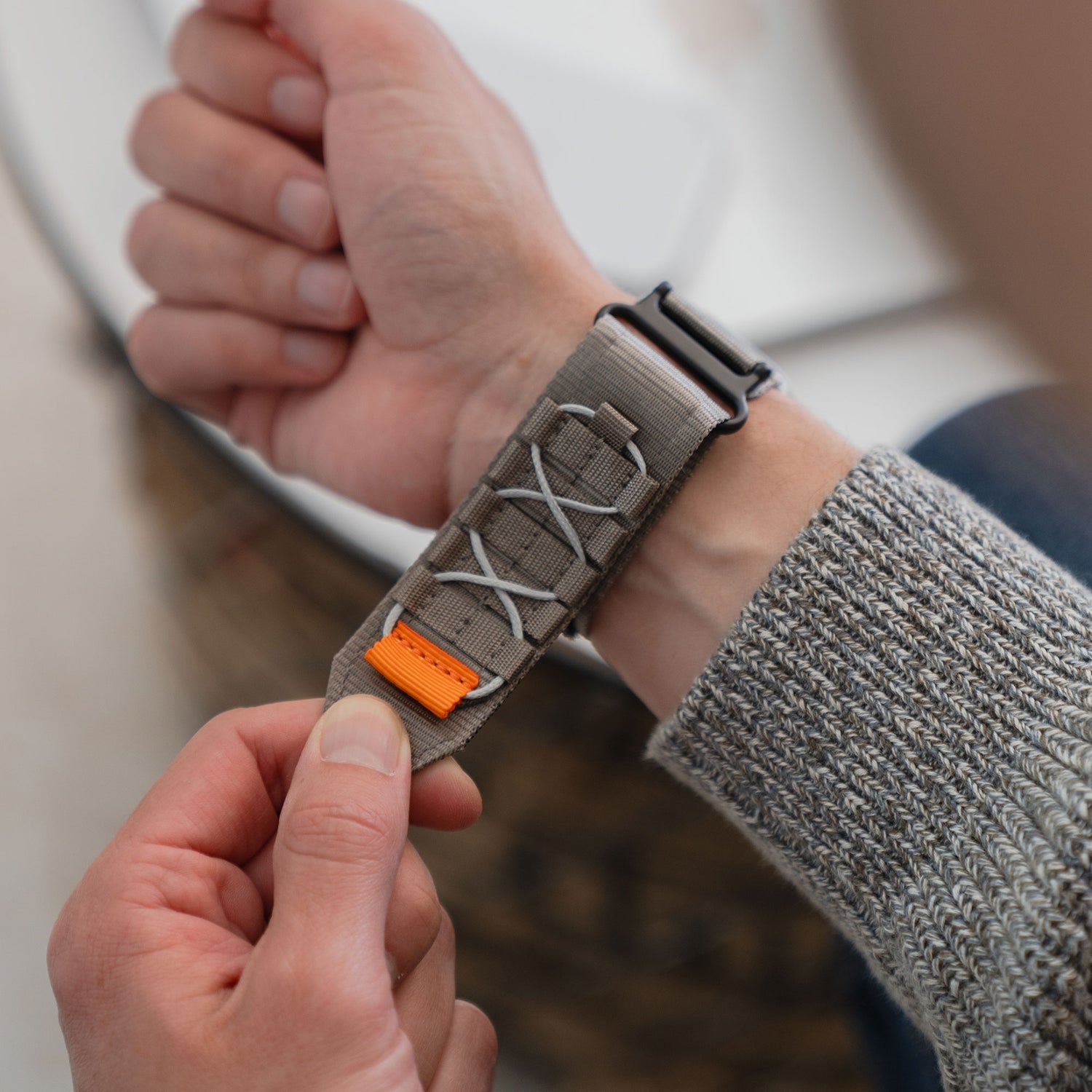 Jemand legt sich das arktisband Apple Watch Outdoor Armband "Forge", ein graues Nylonarmband mit weißen Schnürsenkeln und einem orangefarbenen Akzent von arktisband, um das Handgelenk.