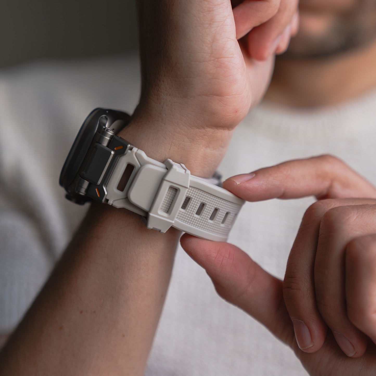 Eine Person befestigt das arktisband Apple Watch Ultra Armband „Atlas“ – ein weißes Band mit robustem Riemen und Edelstahlakzenten von arktisband – am Handgelenk und trägt dabei einen hellen Pullover, auf dem möglicherweise eine Apple Watch Ultra zu sehen ist.