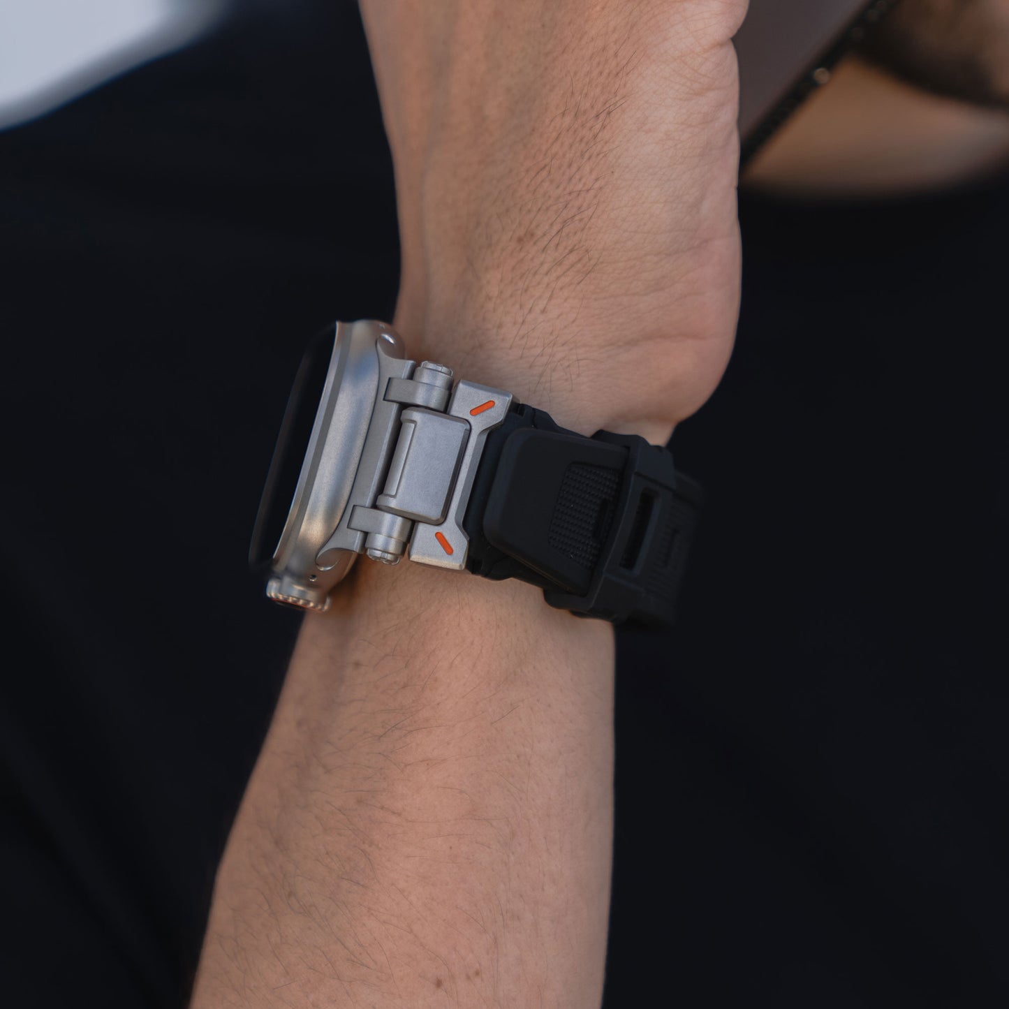 Eine Nahaufnahme einer Person, die ein Arktisband Apple Watch Ultra Armband „Atlas“ mit Edelstahlelementen und orangefarbenem Akzent trägt, ein Smartphone ans Ohr hält und ein schwarzes Hemd trägt.