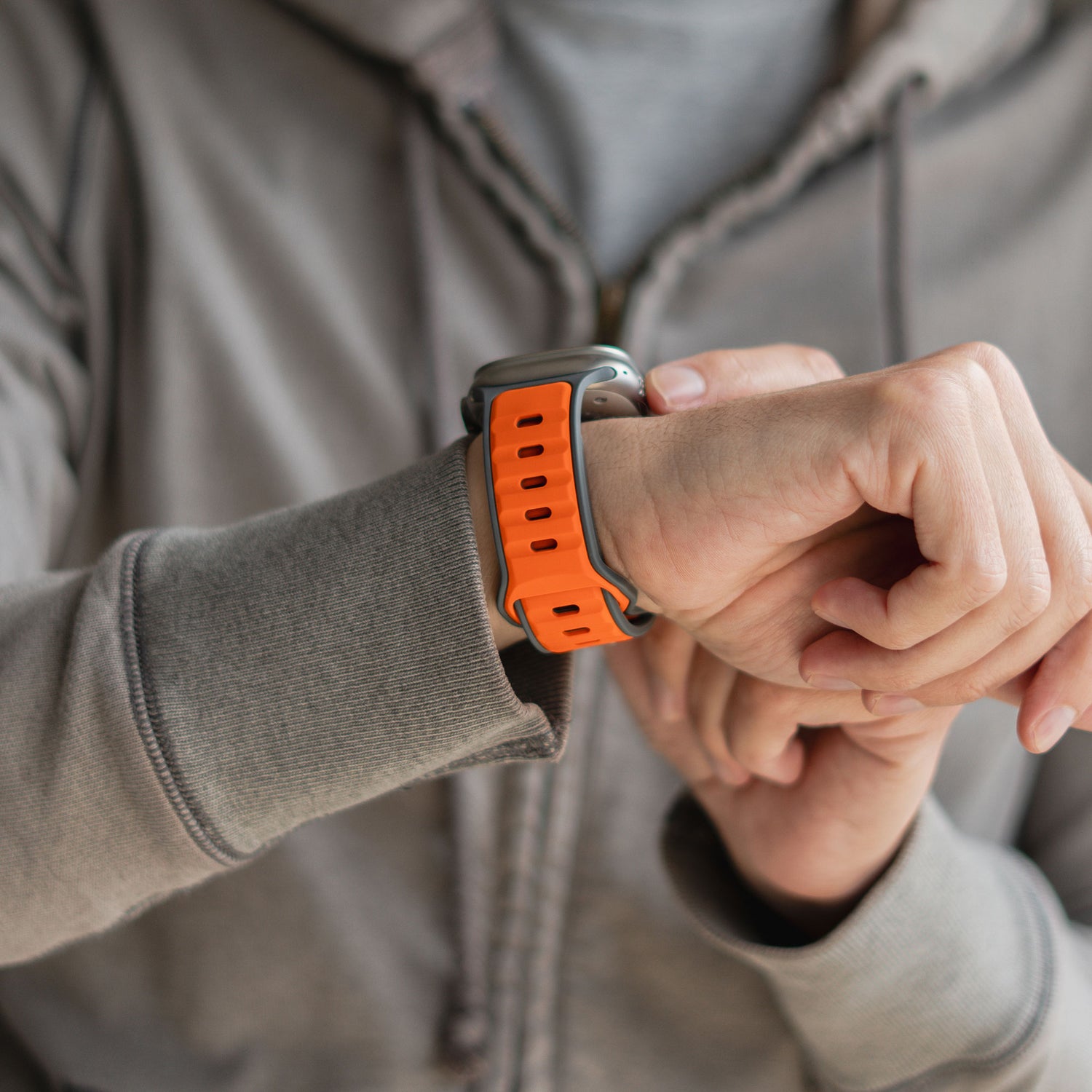 Eine Person in einem grauen Kapuzenpullover checkt ihre Apple Watch. Am linken Handgelenk trägt sie das orangefarbene arktisband „Swift“ Silikonarmband. Im Fokus steht das schweißabsorbierende und atmungsaktive Design von arktisband.