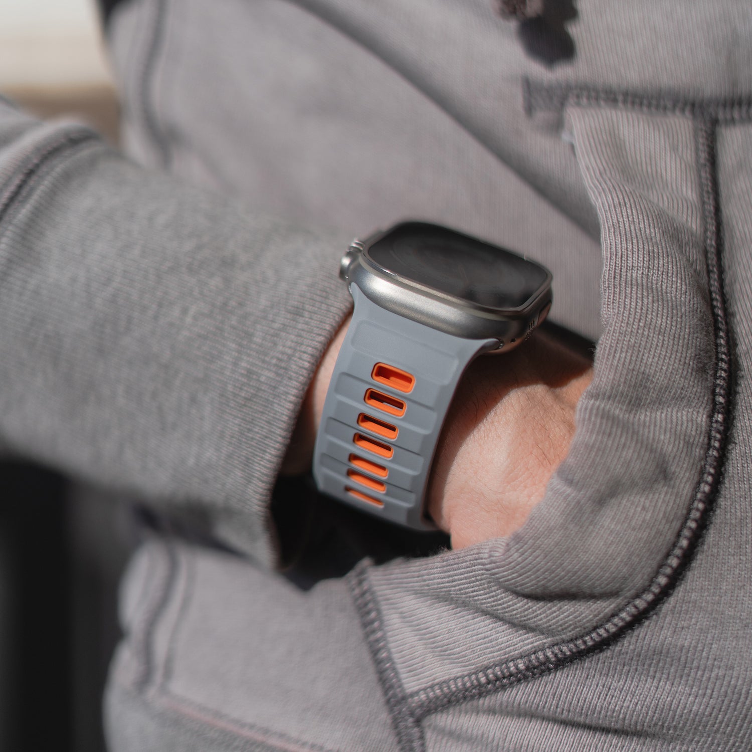 Eine Person in einem grauen Sweatshirt mit der linken Hand in der Tasche trägt ein Arktisband Apple Watch Silikonarmband „Runner“, ein extrabreites, orange akzentuiertes Silikonarmband, das mit der Apple Watch kompatibel ist und über ein dunkles, unbeleuchtetes Display verfügt.