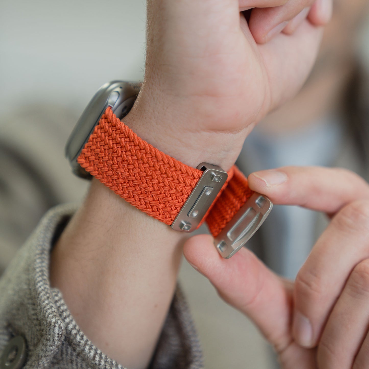 Eine Person befestigt ein orangefarbenes „Arktisband“ – ein geflochtenes Apple Watch Ultra-Armband „Casual“ mit Magnetverschluss – an ihrem Handgelenk und passt dabei den magnetischen Metallverschluss des Arktisband-Armbands an.