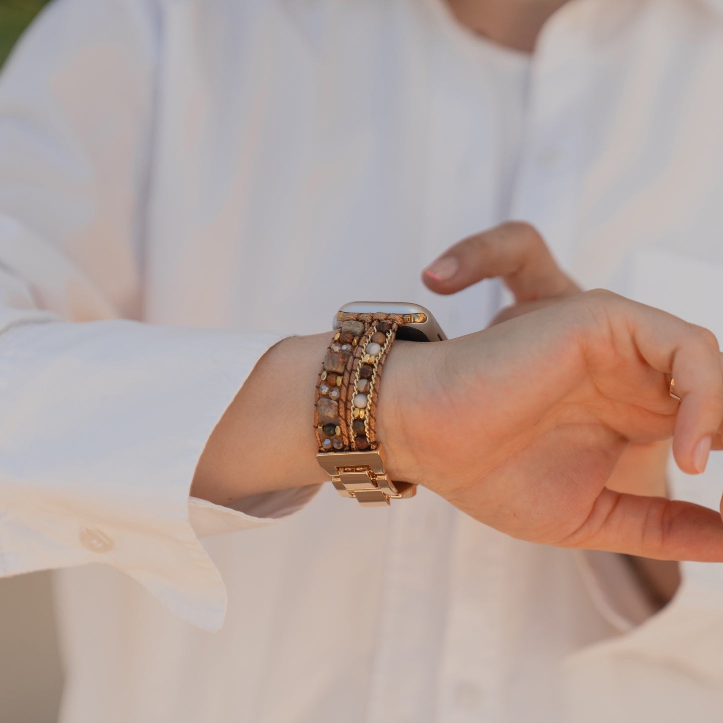 Eine Person in einem weißen Hemd berührt am Handgelenk ein arktisband Apple Watch-Armband „Sunset Spirit“ von arktisband, das mit goldenen und braunen Perlen verziert ist.