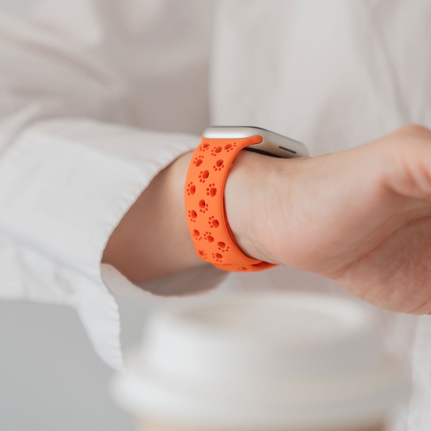 Eine Person in einem weißen Hemd trägt ein orangefarbenes arktisband Apple Watch Silikonarmband "Paw" von arktisband und hält eine Tasse im Hintergrund.
