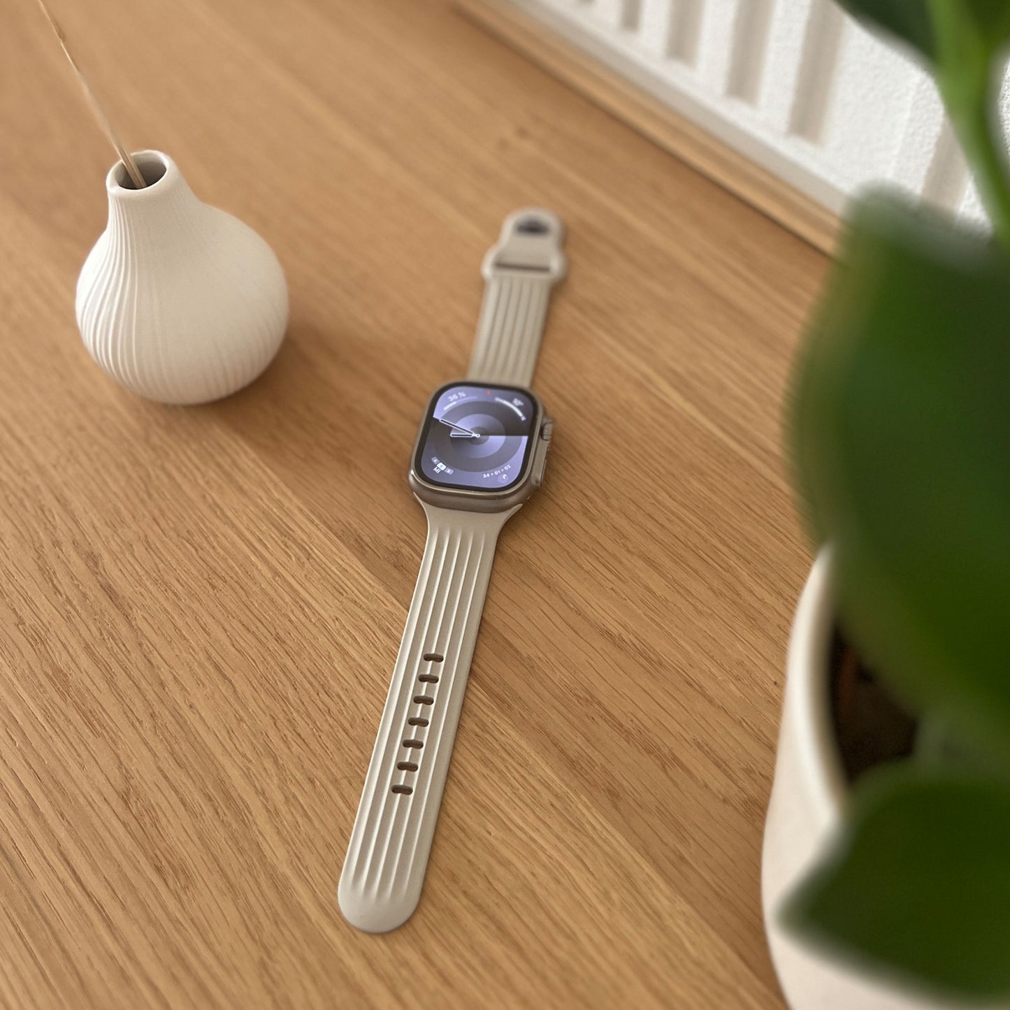 Das arktisband Apple Watch Armband "Skandi" aus beigem Fluorelastomer ruht auf einer Holzfläche neben einer Vase und einer grünen Pflanze und unterstreicht das elegante Skandi-Design der Marke.