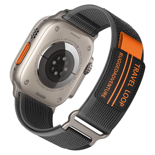 Nahaufnahme eines Arktisband Apple Watch Travel Loop Armbands an einer Apple Watch Ultra mit grauem Gehäuse. Das schwarze Armband hat einen orangefarbenen Streifen, die Aufschrift „TRAVEL LOOP RUGGED ADVENTURE“ und besteht aus strapazierfähiger Nylonfaser. Sensoren und Text sind auf der Rückseite der Uhr sichtbar.