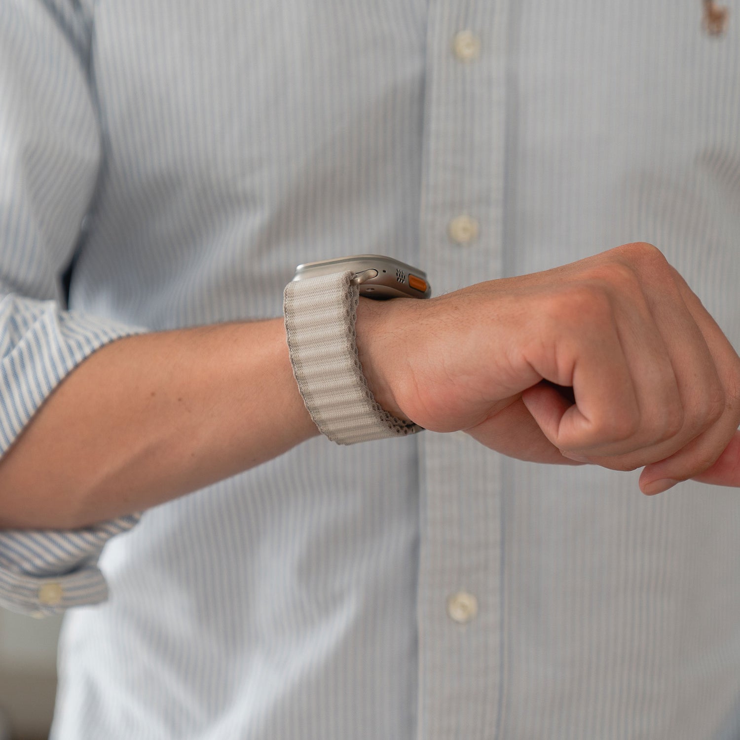 Eine Person in einem hellblauen, gestreiften Hemd mit hochgekrempelten Ärmeln zeigt an ihrem linken Handgelenk das Arktisband Apple Watch Nylonarmband „Venture“ – ein helles, gewebtes Band, ideal für Outdoor-Enthusiasten.