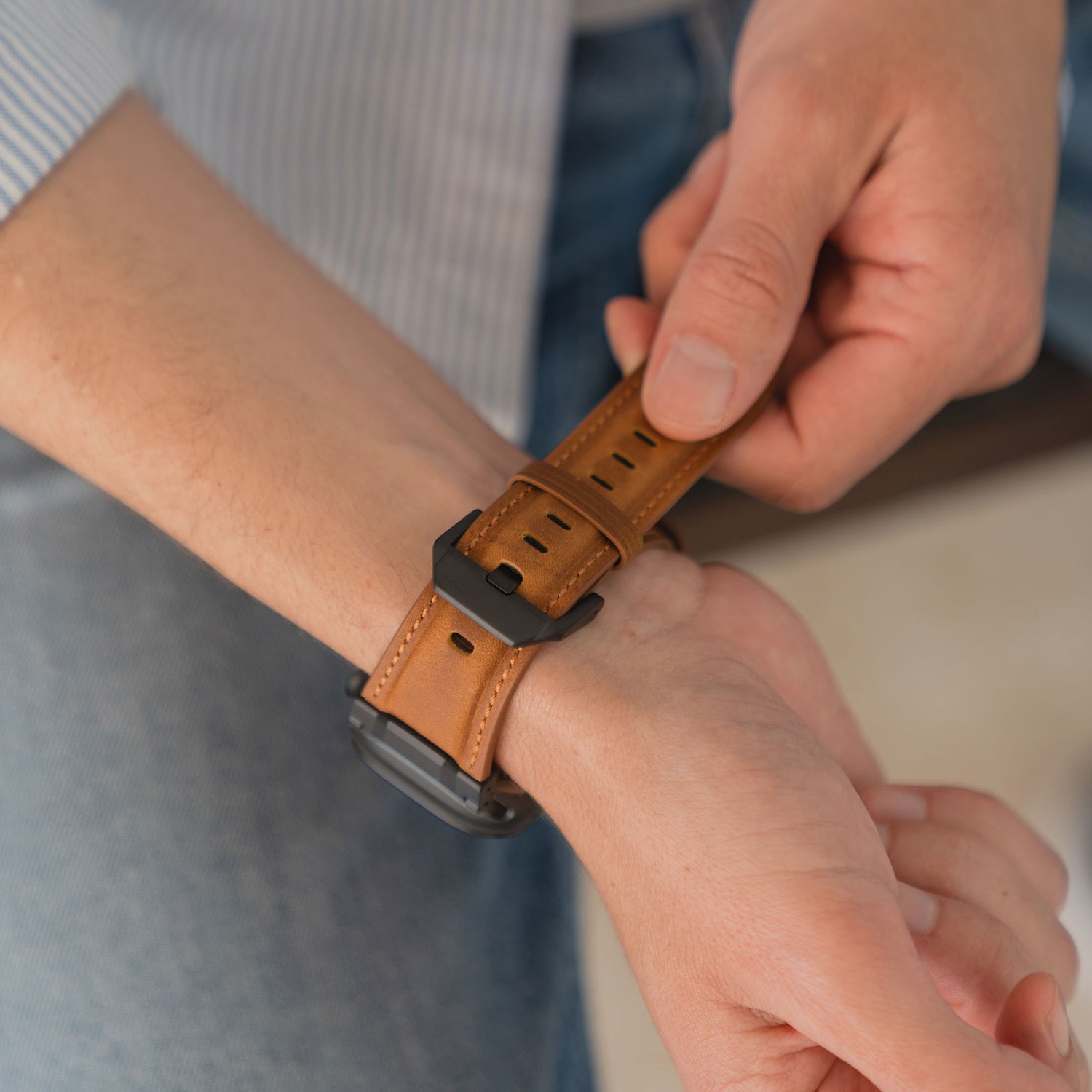 Eine Person legt das arktisband Apple Watch Ultra Lederarmband "Delta" der Marke arktisband an ihr Handgelenk und trägt dazu blaue Jeans sowie ein gestreiftes Shirt.