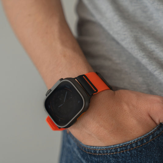 Nahaufnahme einer Person, die das arktisband Apple Watch Armband "Terra" von arktisband, ein extrabreites 24 mm Nylonband in Orange, an ihrer Smartwatch trägt, während sie in Jeans gekleidet ist und eine Hand in der Tasche hat.