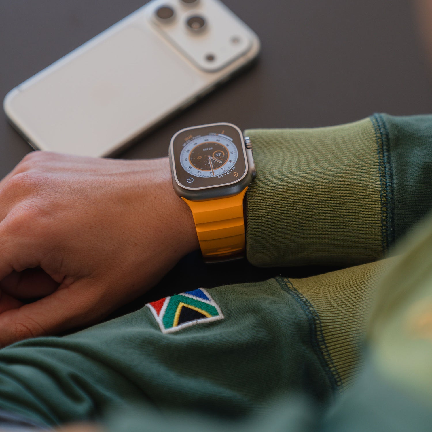 Eine Person, die ein arktisband Apple Watch Armband "Flux" mit Edelstahl Schließe und einem gelben Band trägt, dazu eine grüne Jacke, entspannt sich neben einem weißen Smartphone auf einem Tisch.