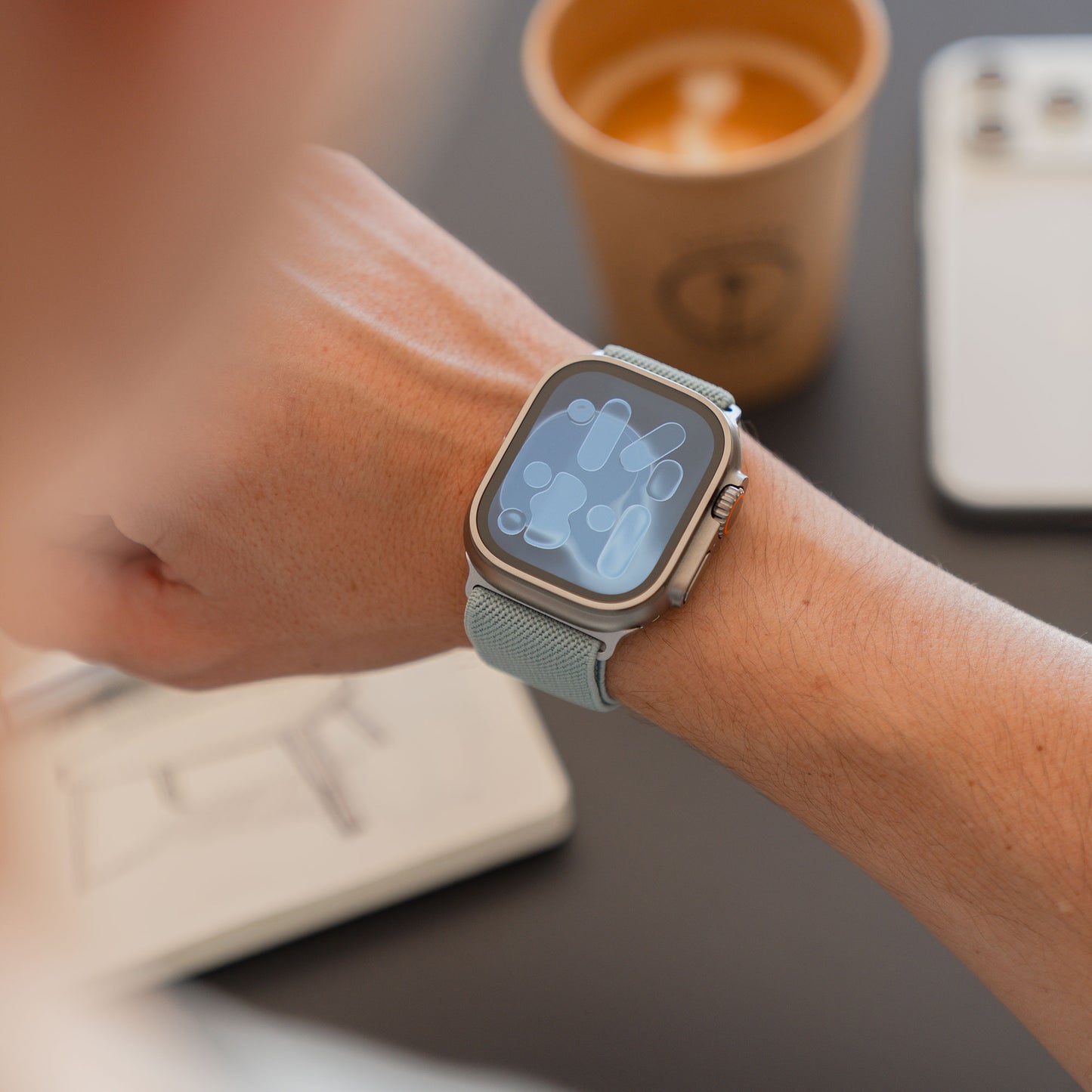 Person, die ein Arktisband Apple Watch Nylonarmband "FlexOne" mit einem abstrakten Display trägt, eine Kaffeetasse hält und ein Telefon und ein Notebook in der Nähe hat.