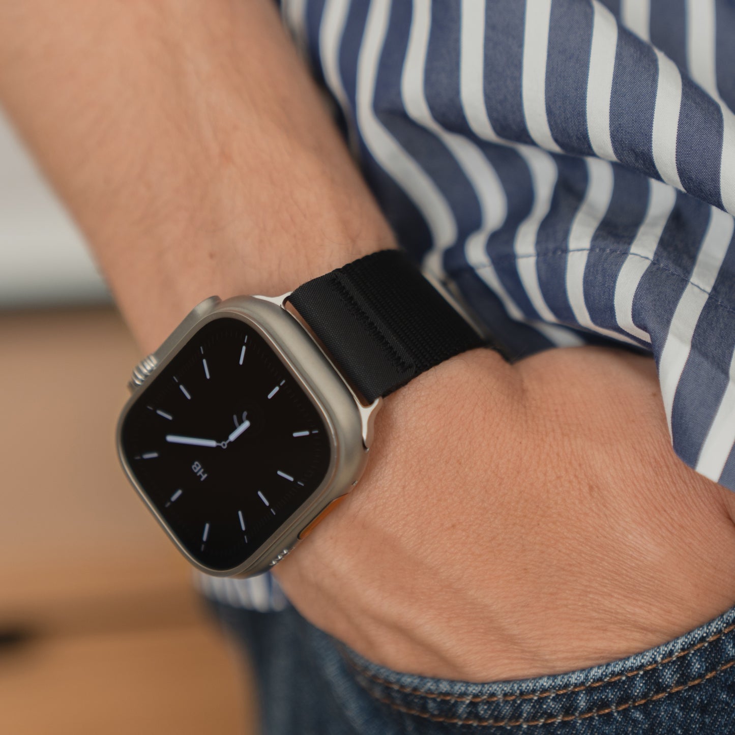 Eine Person trägt ein arktisband Apple Watch Armband "Terra" von arktisband mit einem extra breiten 24 mm Nylonband und ein gestreiftes Hemd, die Hand in der Jeanstasche.