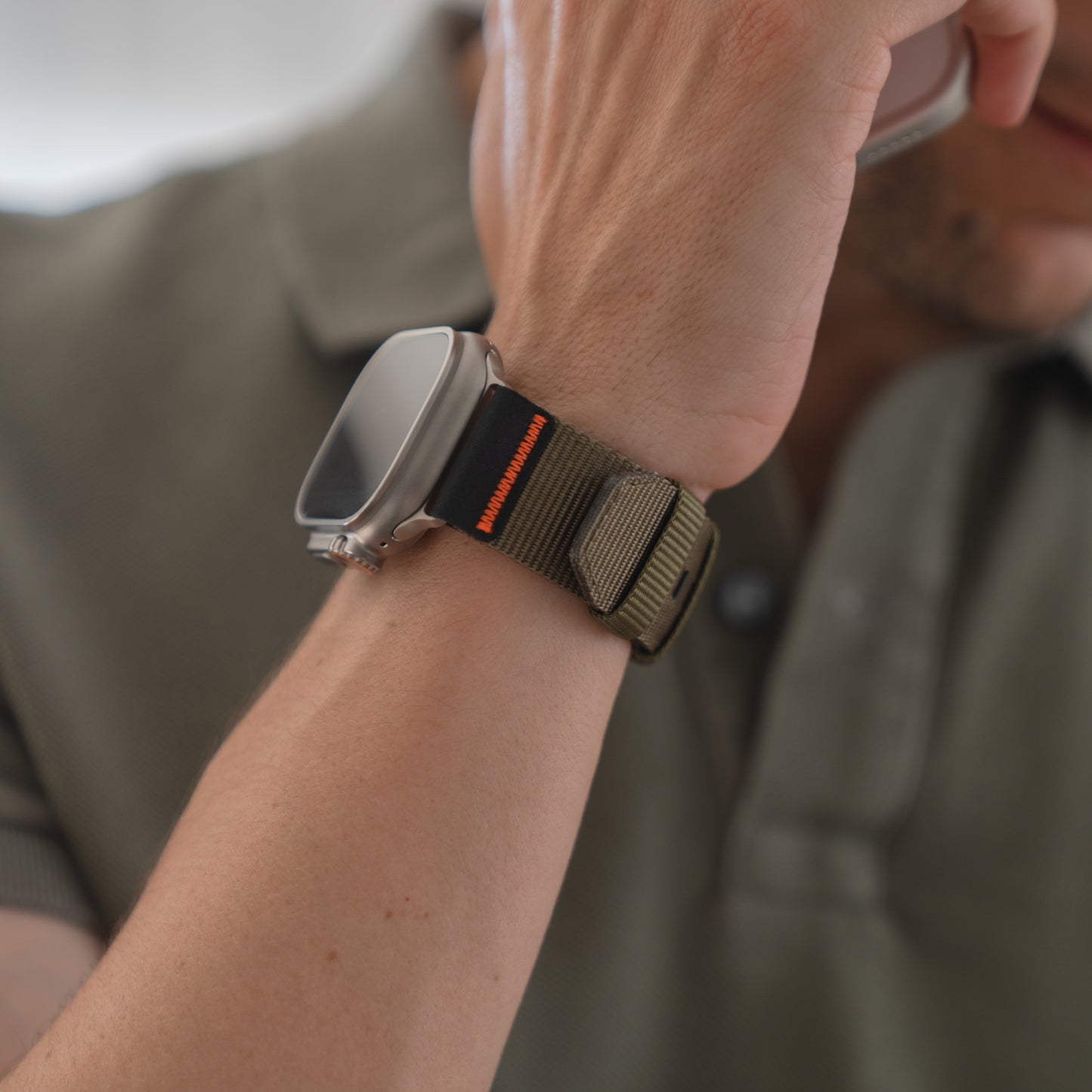 Eine Person in einem grünen Hemd trägt ein arktisband Apple Watch Armband "Terra" von arktisband (extra breites 24 mm Nylonband) an ihrer Apple Watch Ultra und hält ein Telefon ans Ohr.