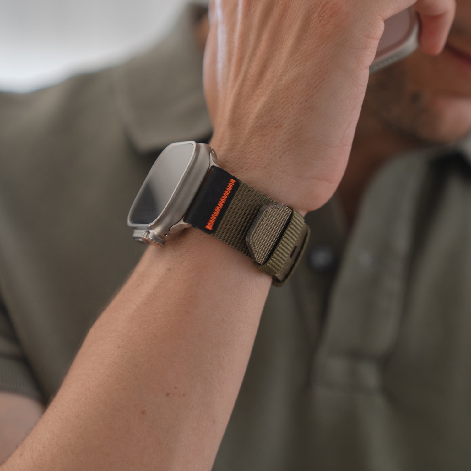 Eine Person in einem grünen Hemd trägt ein arktisband Apple Watch Armband "Terra" von arktisband (extra breites 24 mm Nylonband) an ihrer Apple Watch Ultra und hält ein Telefon ans Ohr.