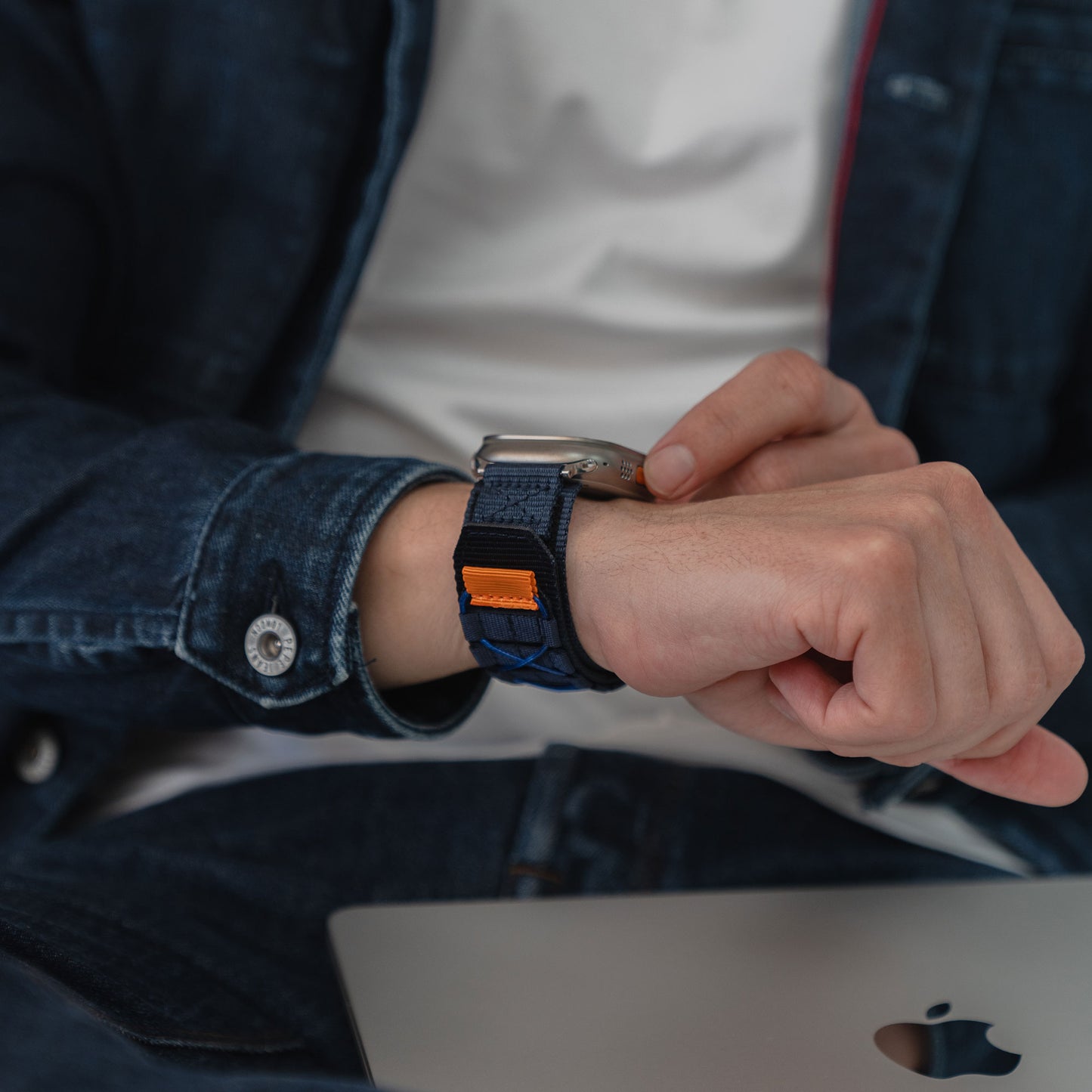 Eine Person in Jeans prüft das arktisband Apple Watch Outdoor Armband "Forge" von arktisband, während sie mit einem Laptop sitzt und einen lässigen Outdoor-Look unterstreicht.
