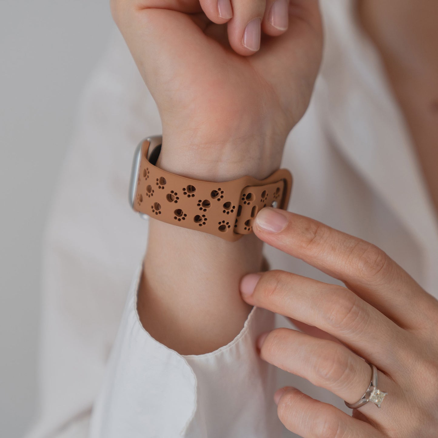 Eine Hand, die ein weißes Hemd und einen Ring trägt, stellt ein beiges Arktisband Apple Watch Silikonarmband "Paw" mit schwarzen Pfotenabdrücken ein.