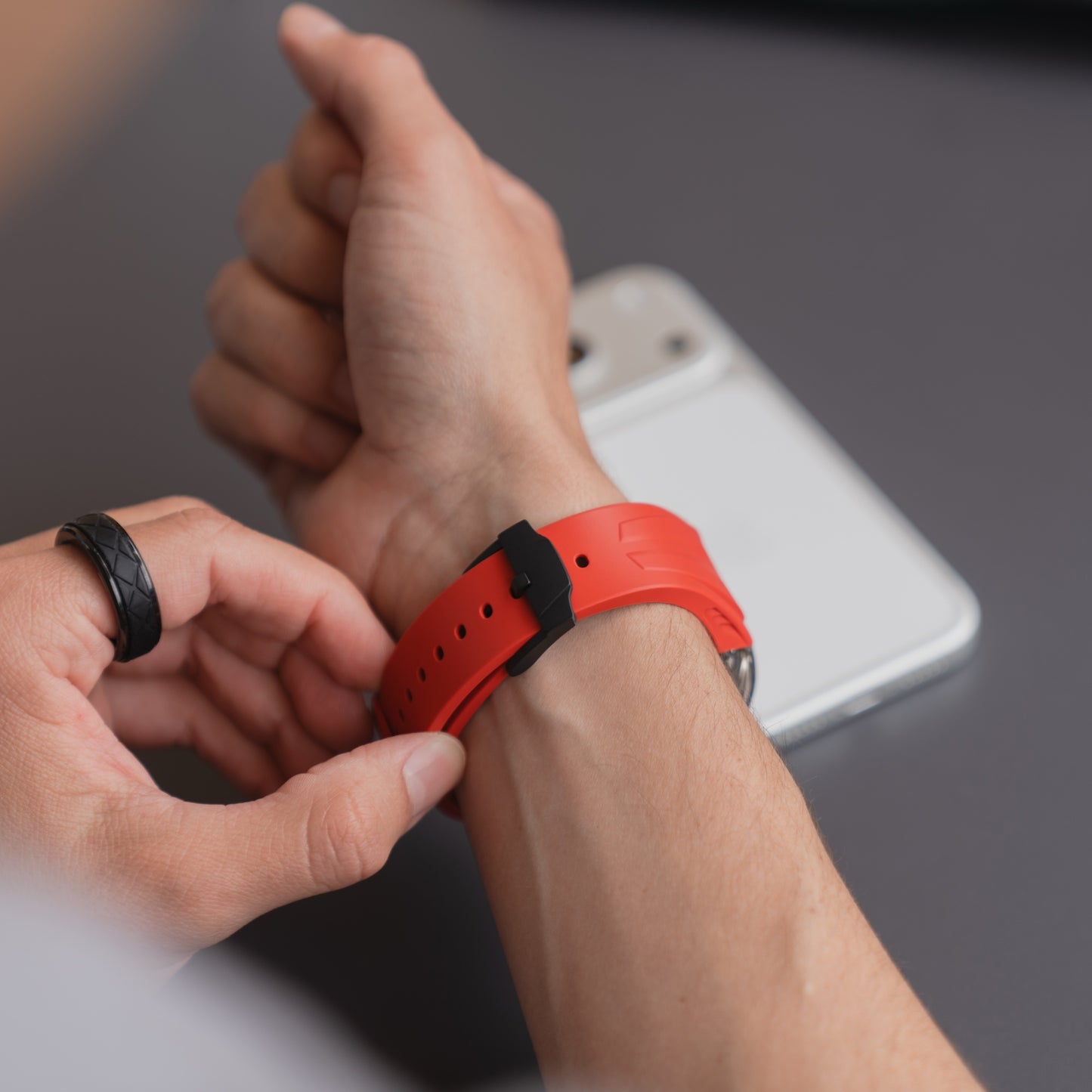 Eine Person trägt das rote arktisband Apple Watch Armband "Armor Fusion" von arktisband am Handgelenk neben einem weißen Smartphone und einem transparenten Hardcase auf grauer Oberfläche.