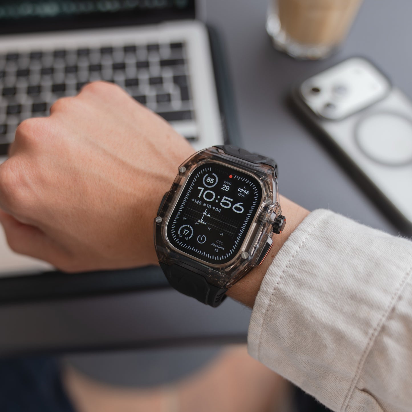 Jemand, der das arktisband Apple Watch Armband "Armor Fusion" in Schwarz trägt, arbeitet an einem Laptop am Schreibtisch, während sein Smartphone in einem transparenten Hardcase liegt.