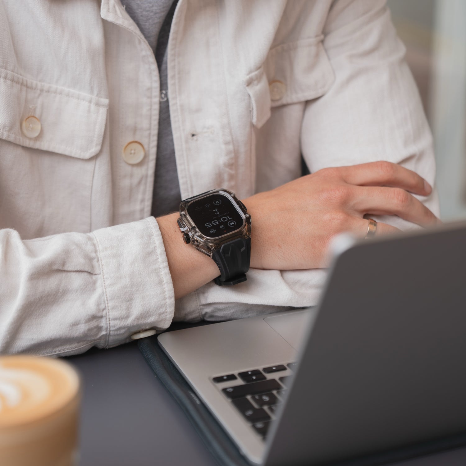 Eine Person in einer weißen Jacke benutzt einen Laptop, während sie das arktisband Apple Watch Armband "Armor Fusion" von arktisband trägt, mit einer Kaffeetasse und einem Hardcase in der Nähe.