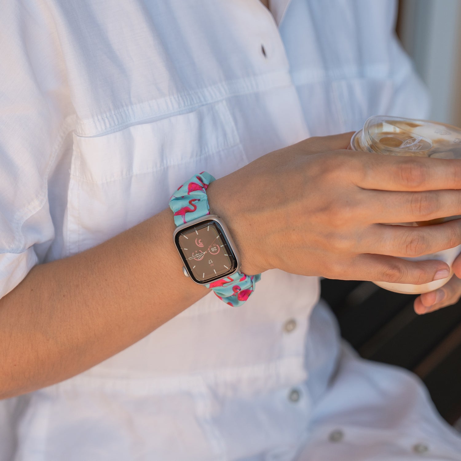 Eine Person in einem weißen Hemd hält ein Getränk in der Hand und trägt das arktisband Apple Watch Scrunchie Armband "Elanora", ein buntes, elastisches Band von arktisband, an ihrem Handgelenk.