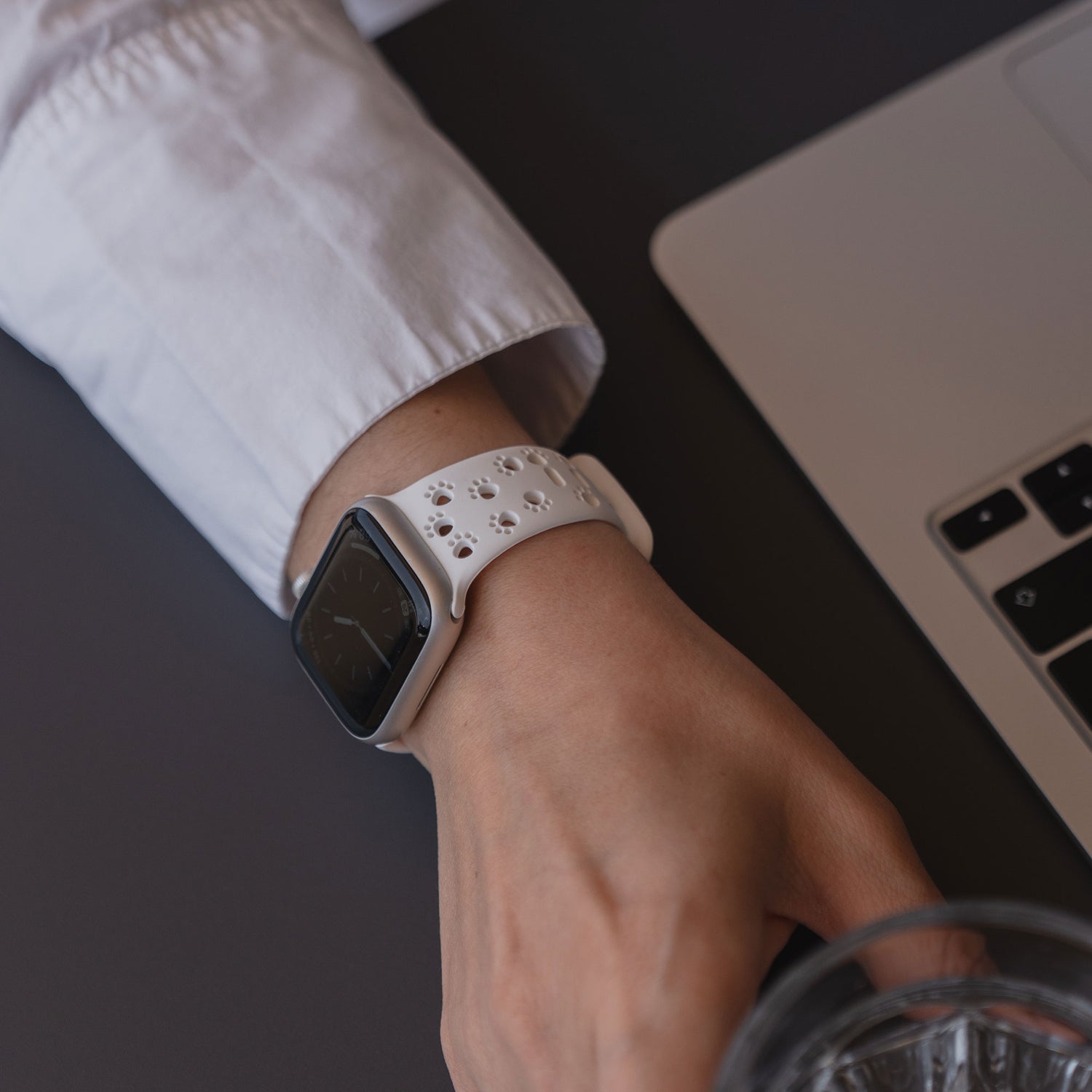 Eine Person, die das arktisband Apple Watch Silikonarmband "Paw" mit weißem Band trägt, greift nach einem Glas in der Nähe eines Laptops, was das minimalistische Pfoten Design von arktisband unterstreicht.