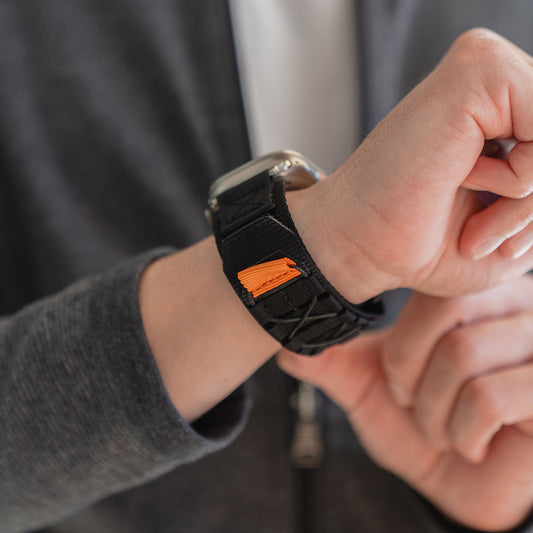 Eine Person passt ein arktisband Apple Watch Outdoor Armband "Forge" mit einem schwarz-orangefarbenen Stoffband an ihr Handgelenk an.