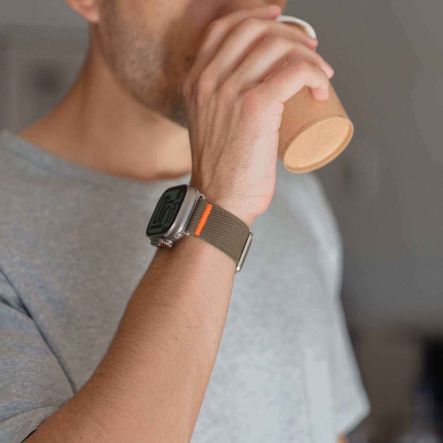 Eine Person in einem grauen Hemd trinkt aus einem Pappbecher und trägt das Arktisband Apple Watch Nylonarmband "FlexOne" mit einem olivgrünen Band.