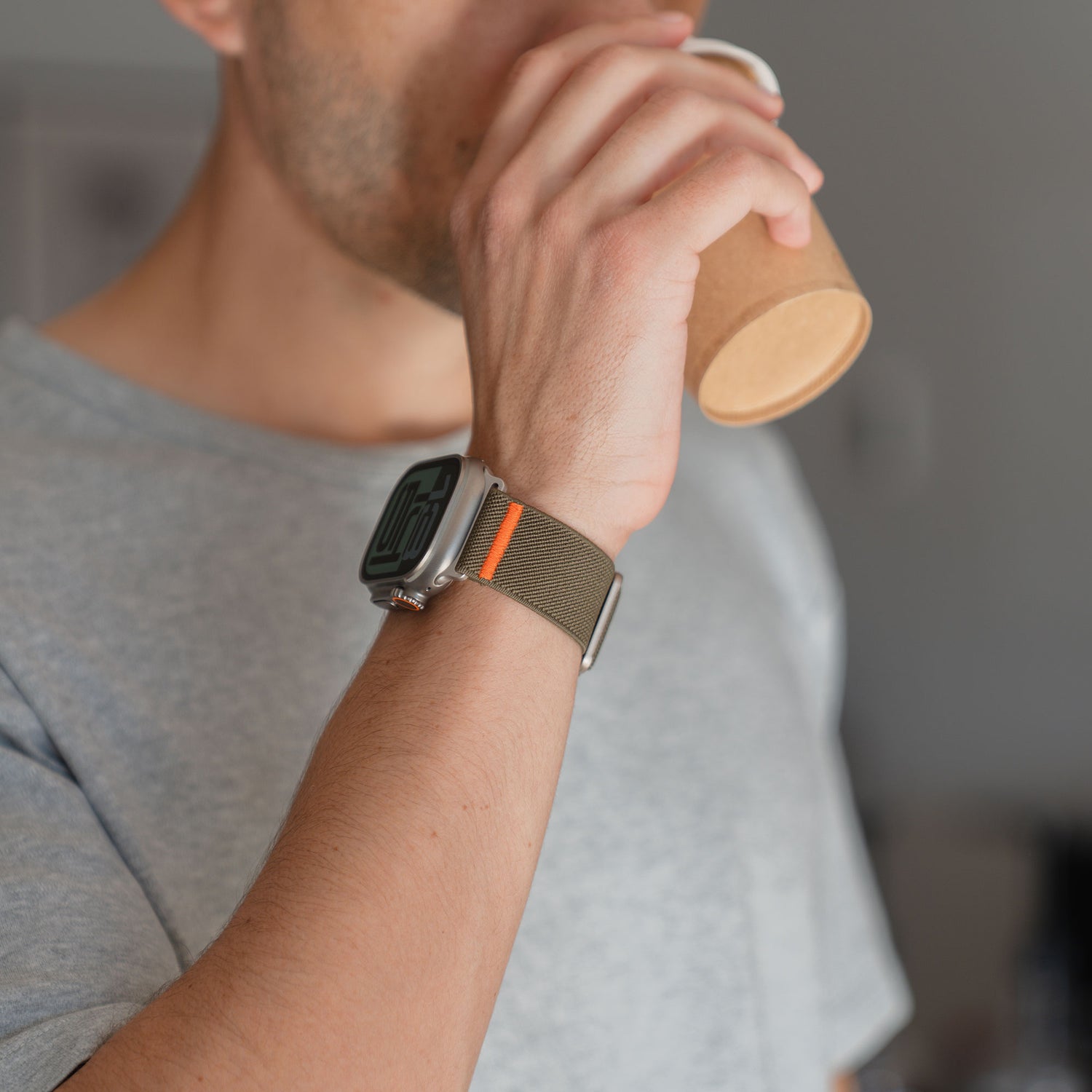 Eine Person in einem grauen Hemd trinkt aus einem Pappbecher und trägt das Arktisband Apple Watch Nylonarmband "FlexOne" mit einem olivgrünen Band.