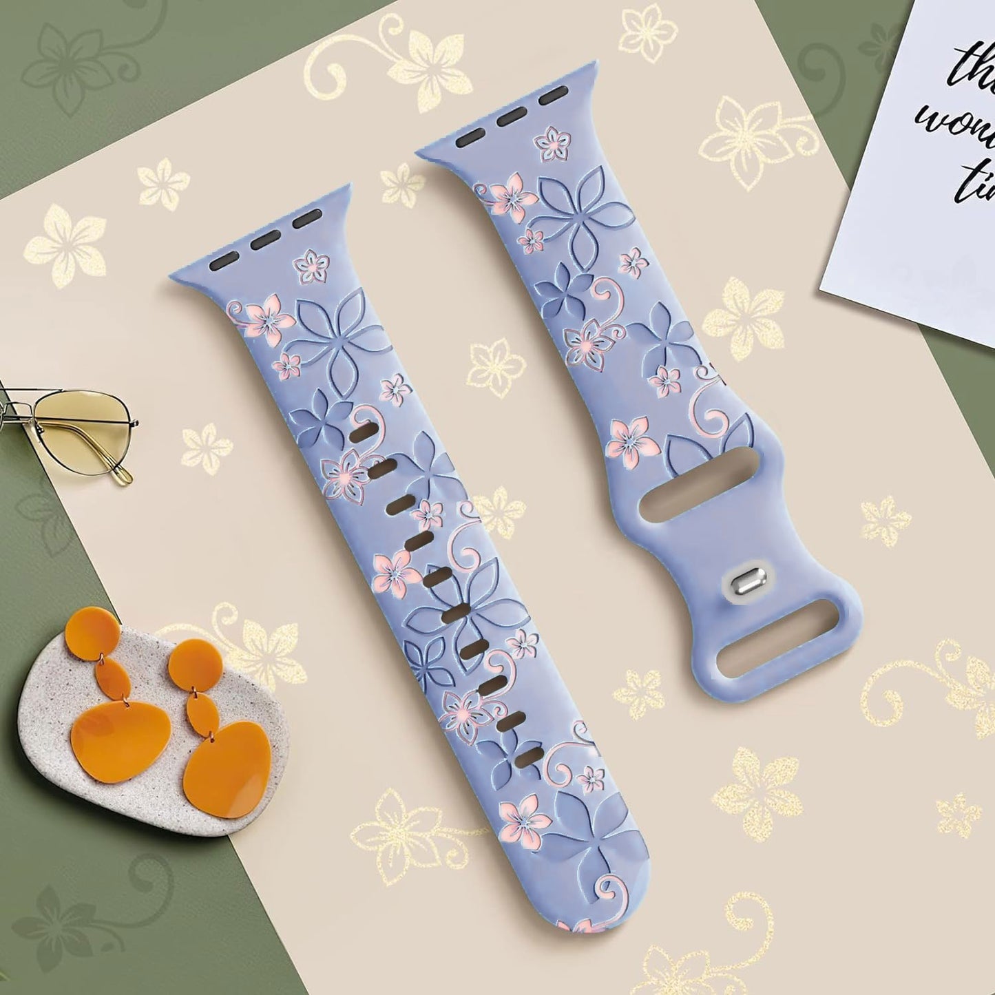 Zwei arktisband Apple Watch Silikonarmbänder "Bloom" mit zartem floralen Muster in Pink und Lila ruhen auf einer gemusterten Oberfläche, umgeben von Schmuck und Brille. Stylische, kompatible Ultra Series, von der Marke arktisband.