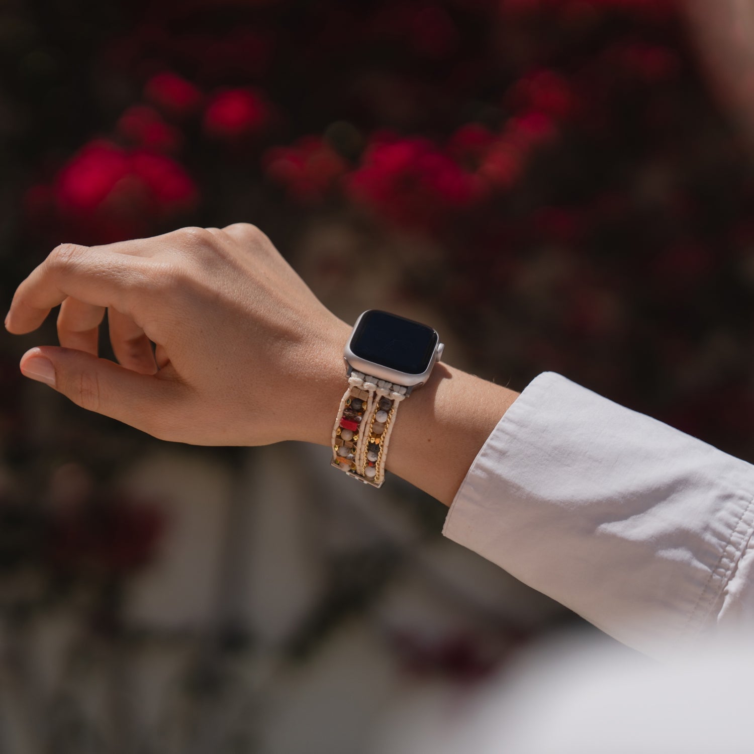 Eine Hand trägt das Arktisband Apple Watch-Armband „Sunset Spirit“ mit Perlenarmband vor einem verschwommenen Hintergrund aus roten Blumen.