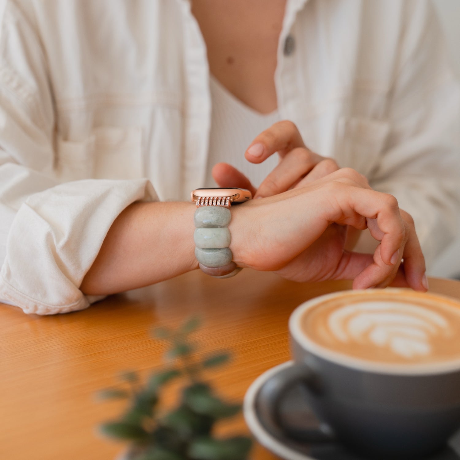 Eine Person in einem weißen Hemd überprüft ihre Apple Watch "Stone Pearl" mit dehnbarem Gummiverschluss, während eine Tasse Latte Art Kaffee auf dem Tisch steht.
