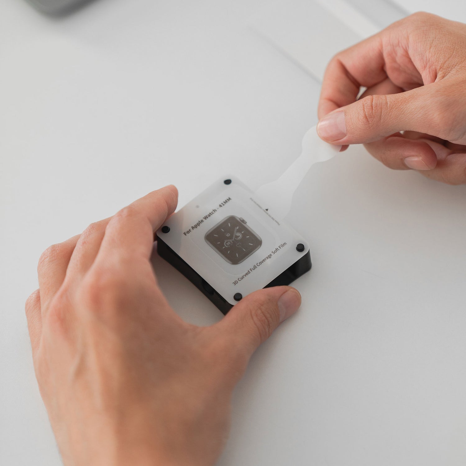 Nahaufnahme von Händen, die eine Arktis SCND.SKIN Apple Watch Full Cover Schutzfolie von einem kleinen elektronischen Gerät auf einer weißen Fläche abziehen.