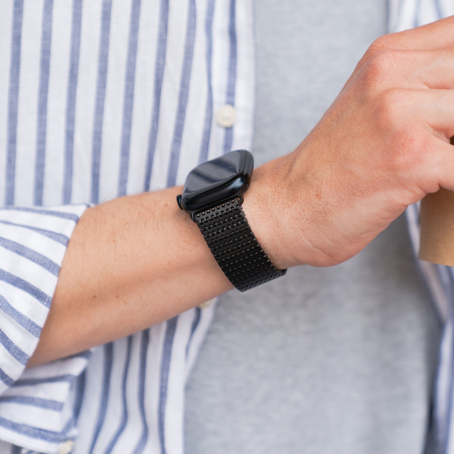 Eine Person, die das Arktisband Apple Watch Edelstahlarmband „Aurora“ trägt, eine Tasse hält und ein gestreiftes Hemd und ein graues T-Shirt trägt.