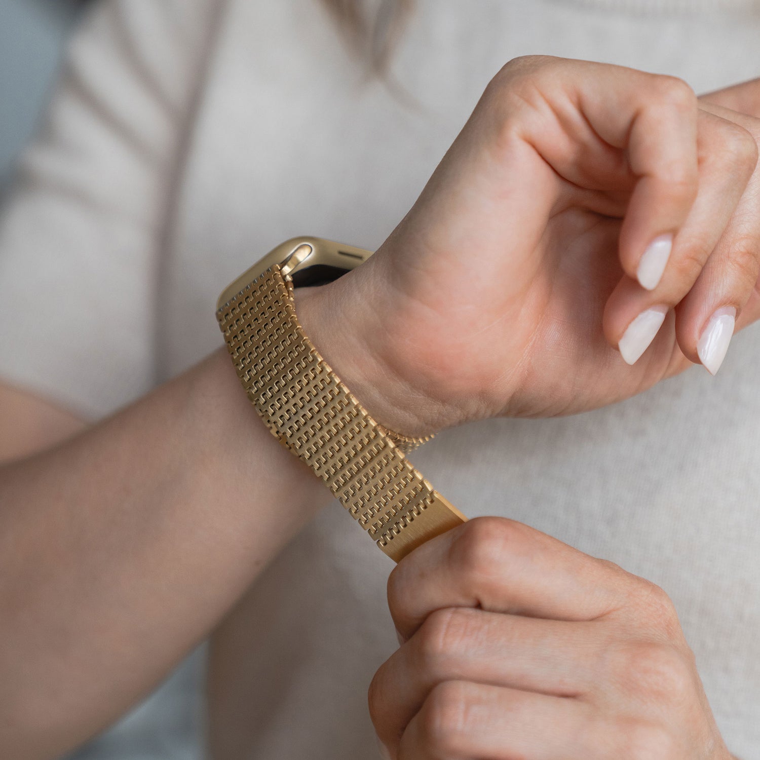 Eine Person befestigt das Arktisband Apple Watch Edelstahlarmband „Aurora“ mit Magnetverschluss und goldenem Mesh am Handgelenk, trägt ein helles Kurzarmoberteil und präsentiert sauber manikürte Nägel.