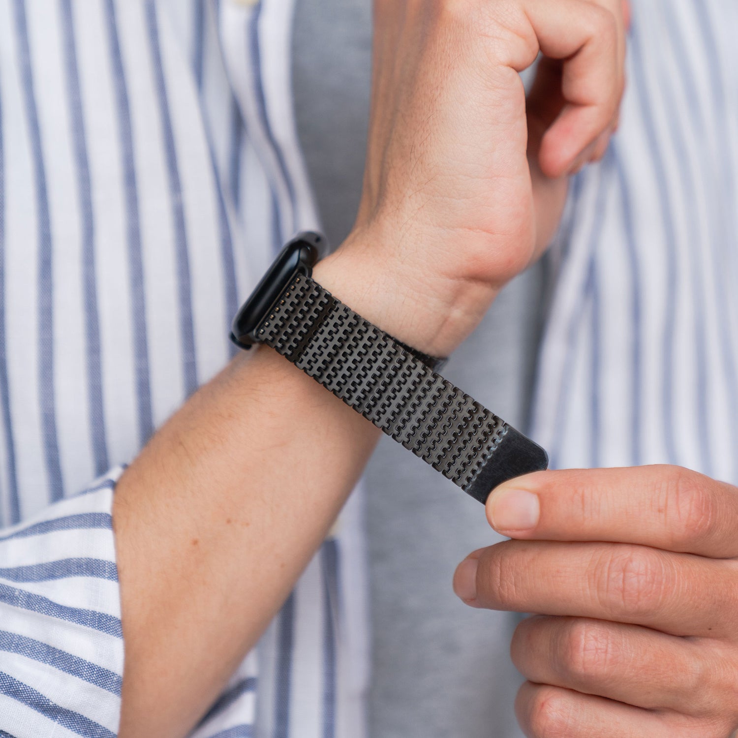 Eine Person befestigt ein schwarzes, strukturiertes Arktisband Apple Watch Edelstahlarmband „Aurora“ mit Magnetverschluss um ihr Handgelenk und trägt dabei ein gestreiftes Hemd.