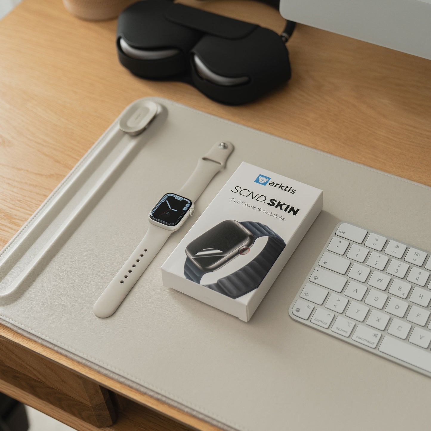 Auf einem aufgeräumten Schreibtisch liegen eine Arktis arktis SCND.SKIN Apple Watch Full Cover Schutzfolie, eine Tastatur, eine Ladestation und eine Coverbox.