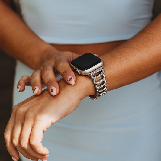 Eine Person in einem hellblauen Oberteil rückt ihre Apple Watch zurecht, die mit dem arktisband „Romance“-Armband ausgestattet ist. Ihre Nägel sind hellrosa lackiert und mit kleinen roten Sternen verziert.