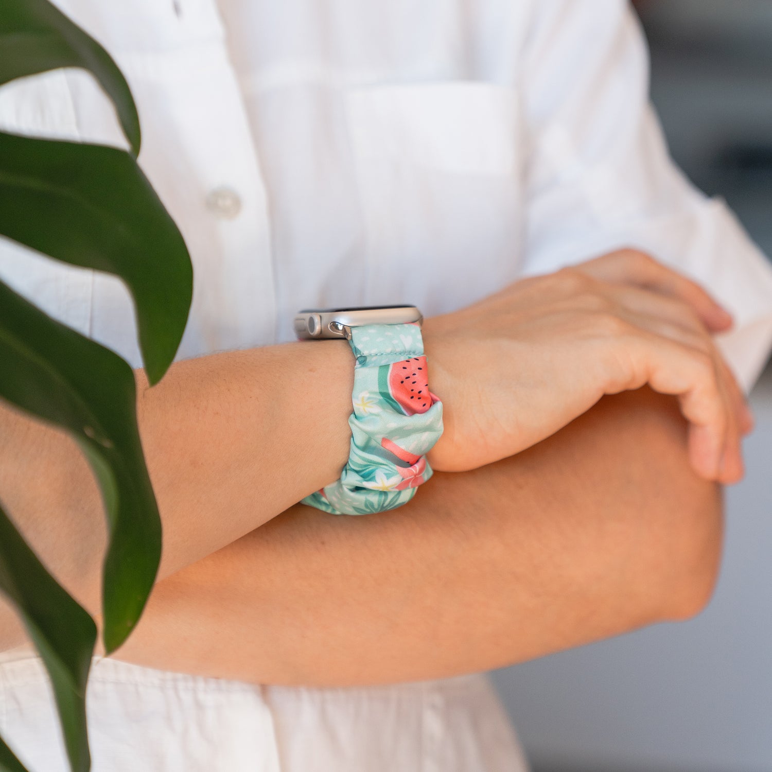 Eine Person trägt das arktisband Apple Watch Scrunchie Armband "Elanora" von arktisband mit einem farbenfrohen, geblümten Gummiband mit einem kleinen Wassermelonen-Anhänger und steht mit verschränkten Armen.