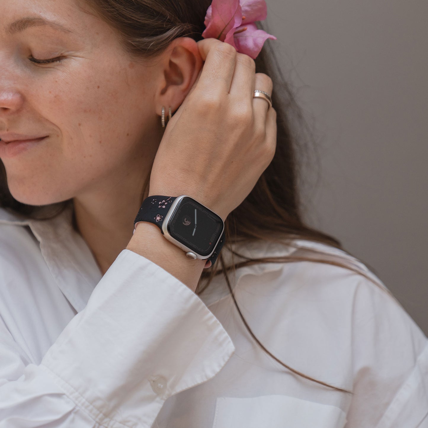 Eine Frau in einem weißen Hemd lächelt und steckt sich eine rosa Blume hinters Ohr, während sie das arktisband Apple Watch Silikonarmband "Bloom" von arktisband mit einem wunderschönen Blumendesign trägt.