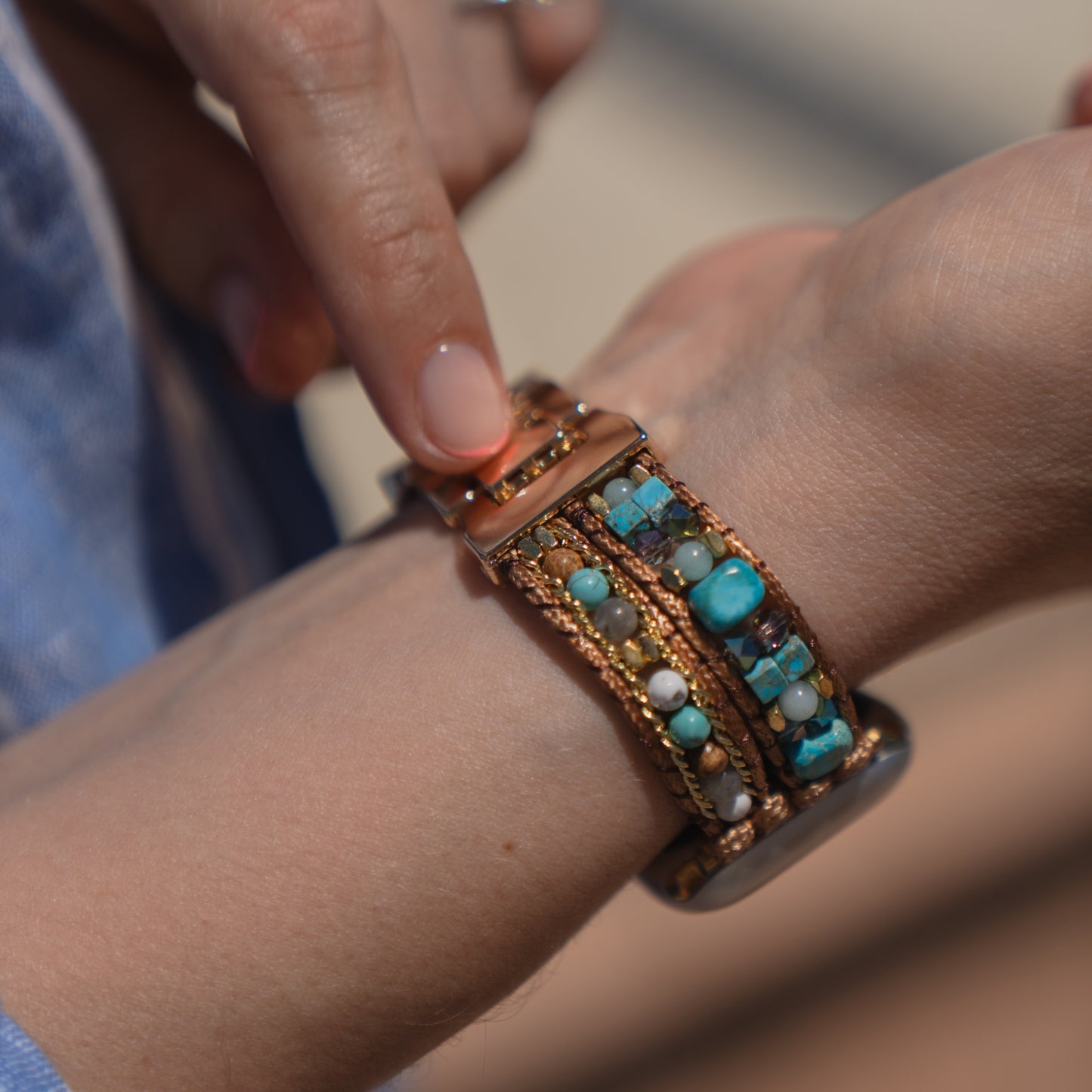 Eine Person legt sich ein „Sunset Spirit“-Armband von arktisband für die Apple Watch mit türkisfarbenen und braunen Perlen am Handgelenk an.