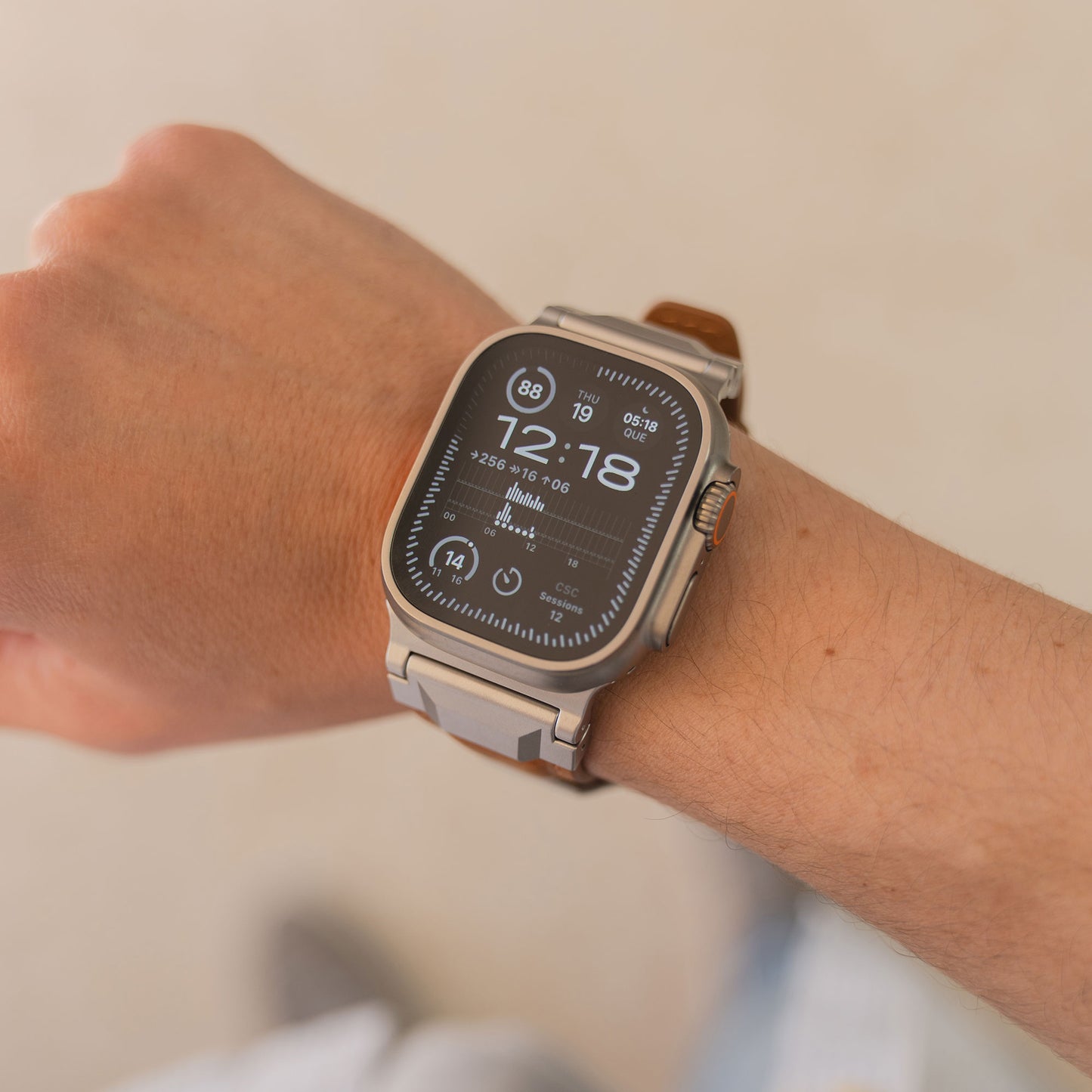 Eine arktisband Apple Watch Ultra mit dem braunen Lederband "Delta" und Edelstahlschließe zeigt 12:18 am Handgelenk einer Person an.
